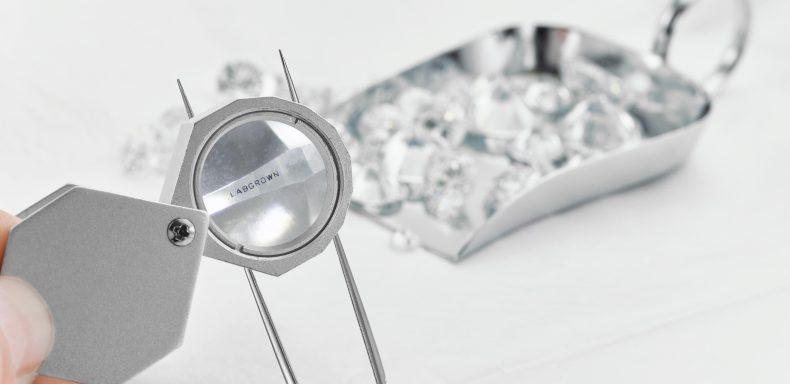 A loupe looking at the words "labgrown" on a piece of paper being held up by a pair of tweezers. In the background is a small tray full of lab diamonds.
